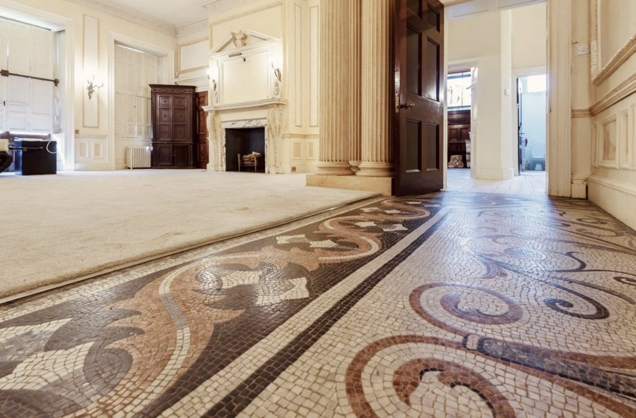 A mosaic floor with scrolling patterns running through a panelled room with a carved fireplace and fluted columns.
