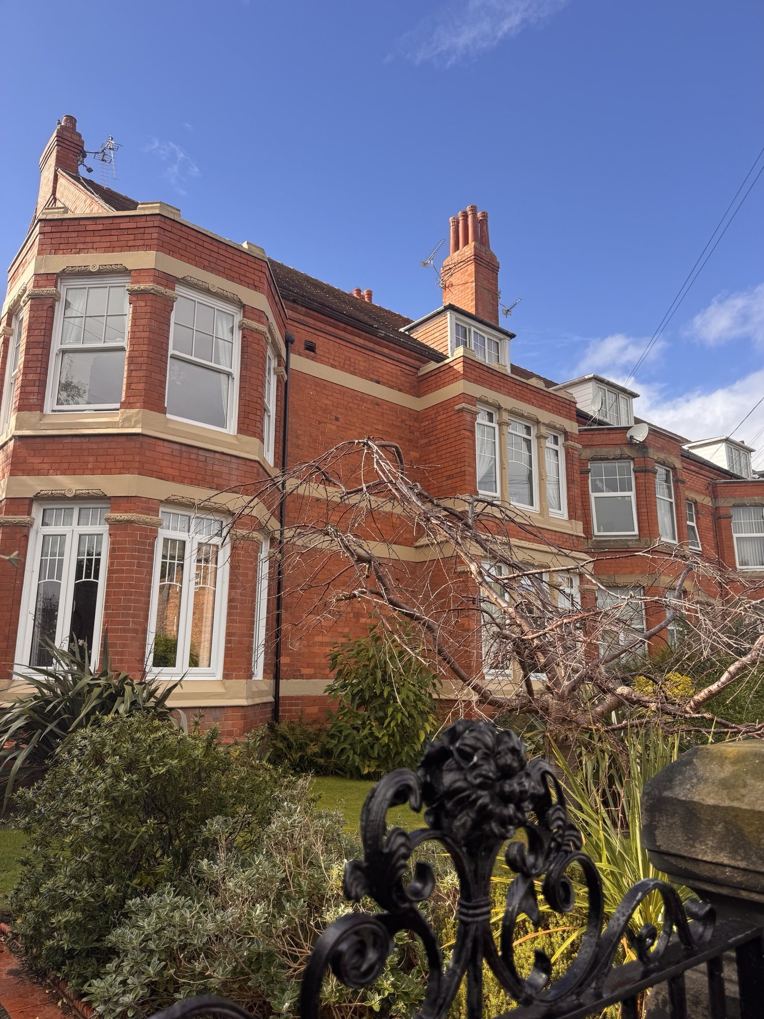 A red-brick house with stone bands, bay windows and tall chimneys, converted into flats, seen over an iron gate.