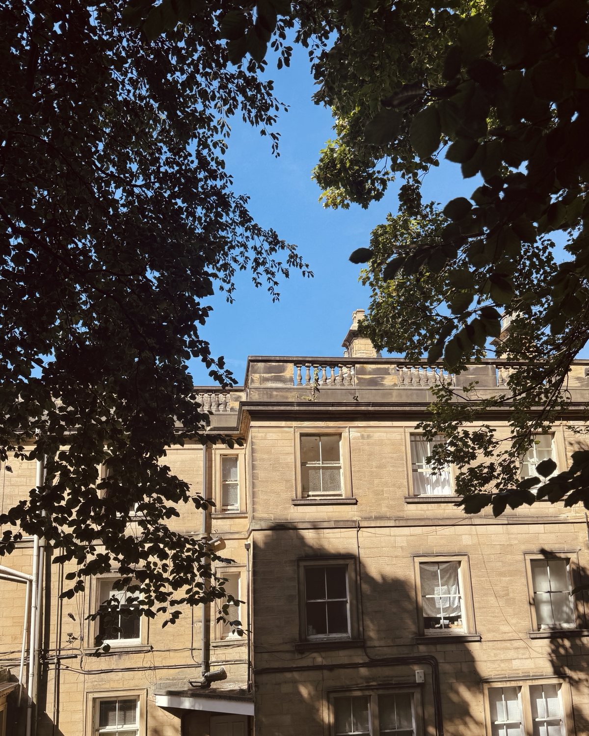 A stone house with a balustrade along the roofline, seen through trees.